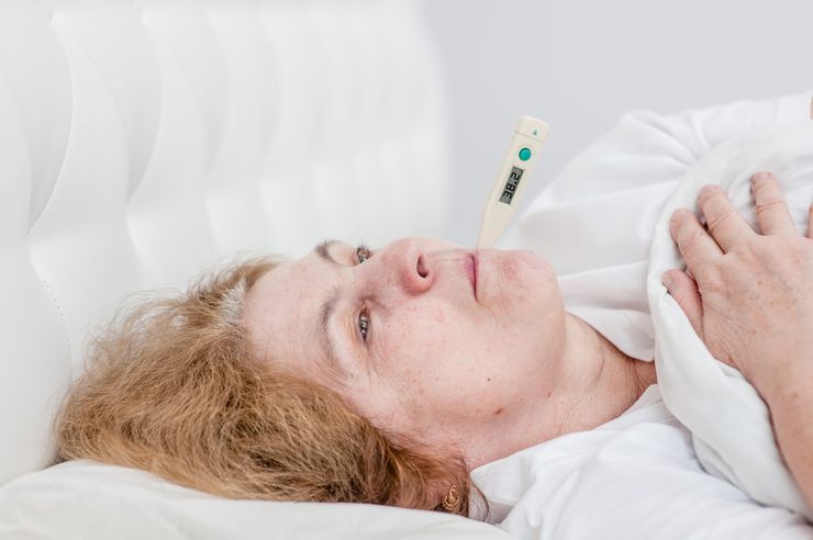 An older woman sick in bed with a digital thermometer showing a fever of 38.2 degrees Celsius during a gout flare
